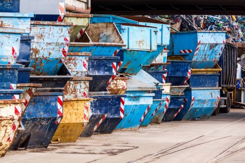 A row of large metal skips and containers used for rubbish and waste collection are lined up on a paved area, with some showing signs of rust and wear. The skips are predominantly blue, with a few black and yellow ones interspersed, and feature red and white reflective safety stripes along their edges. The containers are positioned closely together, some stacked slightly at angles, indicating they are ready for waste disposal or collection services. The surface beneath them is a concrete or asphalt pavement with streaks and minor stains, suggesting frequent use in a waste management setting. In the background, a black waste collection truck is partially visible, emphasizing the commercial rubbish removal context. This scene likely depicts a waste management yard or a storage area for rubbish collection services near Forest Gate, with clear visibility of environmental surroundings typical for such facilities. Commercial Waste Forest Gate offers expert rubbish removal services within this operational environment, supporting local waste disposal needs in the Forest Gate area.