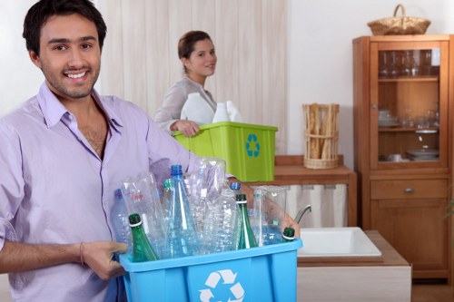 A young man with dark hair and a beard, smiling and holding a blue recycling bin filled with empty plastic bottles, including clear and green-tinted bottles, in a domestic kitchen setting. Behind him, a woman with dark hair tied back, wearing a light grey top, is standing near a green recycling bin with a recycling symbol and a white bag inside. The kitchen features wooden cabinets with glass doors, a white sink, and a wooden countertop. The overall environment is bright and clean, with natural lighting, and the scene emphasizes waste separation and recycling practices relevant to rubbish removal services in Forest Gate, London, focusing on proper disposal of plastic waste.