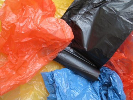 A collection of various large plastic rubbish bags in different colours, including bright orange, black, blue, and yellow, are arranged on a paved surface. The orange and yellow bags are crumpled, while the black bag appears tightly packed and has a smooth finish. The blue bag is partially opened with some material visible at the edges. The bags are likely made of durable plastic and are intended for waste disposal, consistent with rubbish removal services such as those provided by Commercial Waste Forest Gate. The background shows a flat outdoor area, indicating the bags are placed outside, possibly on a driveway or pavement in Forest Gate. The lighting is natural, highlighting the glossy texture of the plastic bags and emphasizing their creases and folds, reflecting common rubbish collection conditions in the local area. This image effectively illustrates typical waste collection materials used in commercial or residential rubbish removal within the Forest Gate locale.