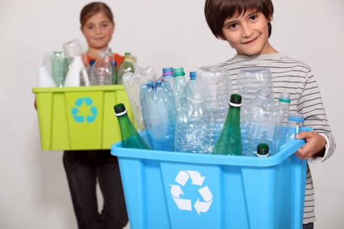 A young boy with dark hair, wearing a striped long-sleeve shirt, stands in the foreground holding a blue recycling bin filled with clear plastic bottles and containers of various sizes, some with green and blue caps. Behind him, a young girl with light brown hair, dressed in a dark long-sleeve top, holds a bright yellow recycling bin containing similar plastic waste. The background is plain and neutral, emphasizing the focus on the waste containers and the children. The scene illustrates the importance of recycling household plastics, relevant to local rubbish removal services offered by Commercial Waste Forest Gate near London, with the setting suggesting an indoor environment suitable for waste sorting and collection.
