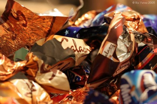 A close-up view of a pile of crumpled foil chocolate wrappers in various metallic colors including copper, silver, and gold, with some featuring printed branding and decorative elements. The wrappers are stacked chaotically, with some partially unfolded or wrinkled, revealing different textures and finishes. The background suggests an indoor setting, possibly on a surface such as a table or countertop, with soft lighting highlighting the metallic sheen of the wrappers. This image illustrates typical waste material that might be collected for rubbish removal services, such as those provided by Commercial Waste Forest Gate, supporting efficient disposal of recyclable packaging waste in the Forest Gate area within the London postcode district.