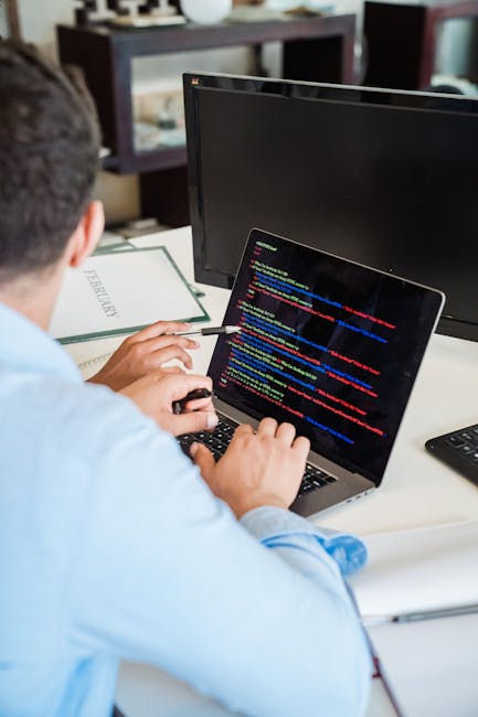 A person seated at a desk, viewed from behind, working on a laptop with a screen displaying colourful lines of code in programming language. The individual, wearing a light blue shirt, is using a mous